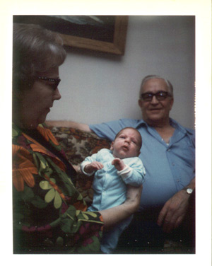 a very 1970s photo showing grandparents in loud floral prints on a loud floral couch holding baby Tommy