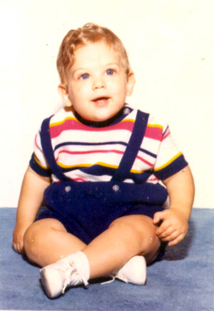 photo of a very chubby Tommy around his first birthday, posed, wearing overall shorts with a striped shirt